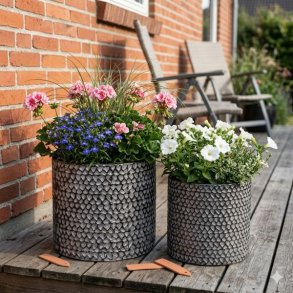 Et stemningsfuldt milj�billede i 1:1 format, der viser de to m�nstrede metalkrukker fra image_98.png og image_99.png, placeret sammen som en gruppe p� et tr�gulv p� en terrasse. Den store krukke (H26,5 cm) og den lille krukke (H23,5 cm) er beplantet med blomstrende pelargonier og petunier, samt dekorativt prydgr�s. Baggrunden viser en r�d murstensv�g og sl�ret havevegetation i sollys.