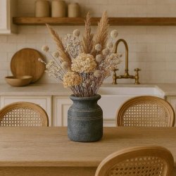 Den rustikke cementkrukke er placeret p� et spisebord af tr� i et lyst k�kken og er fyldt med en stor buket af t�rrede blomster i beige nuancer. I baggrunden ses hvide k�kkenelementer, en messingvandhane og fletstole.