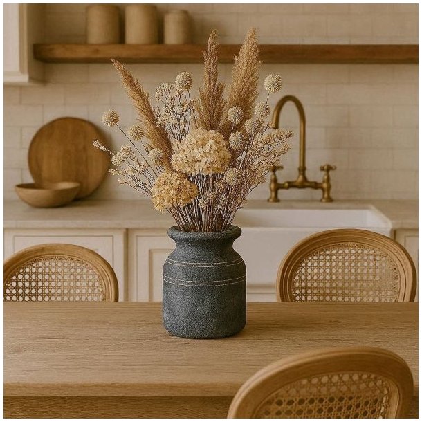 Den rustikke cementkrukke er placeret p� et spisebord af tr� i et lyst k�kken og er fyldt med en stor buket af t�rrede blomster i beige nuancer. I baggrunden ses hvide k�kkenelementer, en messingvandhane og fletstole.