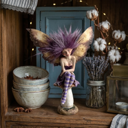Et t�t, stemningsfuldt, kvadratisk milj�billede taget i en lys stue. Den siddende Fairy-figur i polyresin med dramatisk lilla h�r, plettede vinger og stribede ben sidder roligt p� en stor paddehat, der er placeret p� en smuk, patineret tr�hylde. Bagved er der en hyggelig krog med en stack af ceramic sk�le, t�rret lavendel og en lille vase med bomuldsblomster, der fremh�ver feens detaljerede finish og farver.