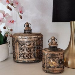 To m�nstrede metallanterner i antik brunguld med t�ndte lys, opstillet p� en sk�nk ved siden af en hvid orkid� og en messinglampe.