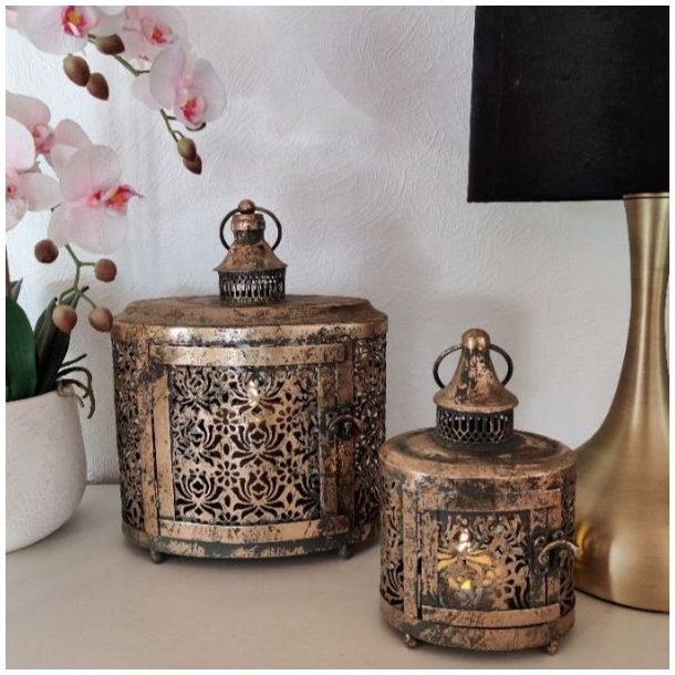 To m�nstrede metallanterner i antik brunguld med t�ndte lys, opstillet p� en sk�nk ved siden af en hvid orkid� og en messinglampe.