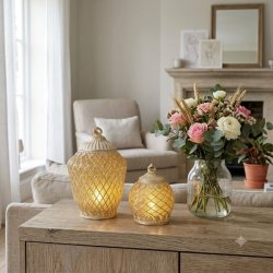 To dekorative, m�nstrede lanterner i en varm guld- og sandfarvet nuance med t�ndt lys indeni, placeret p� en tr�sk�nk i en hyggelig stue ved siden af en glasvase med en flot blomsterbuket.