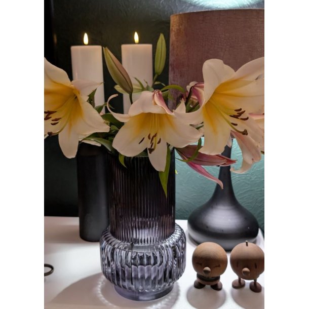 N�rbillede af en m�rk rillet glasvase i retrodesign fyldt med liljer, st�ende i hyggelig belysning ved siden af levende lys.