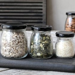 Opbevaringsglas med sorte l�g fyldt med solsikkefr�, gr�skarkerner og sesamfr� p� en m�rk tr�bakke.