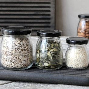 Opbevaringsglas med sorte l�g fyldt med solsikkefr�, gr�skarkerner og sesamfr� p� en m�rk tr�bakke.