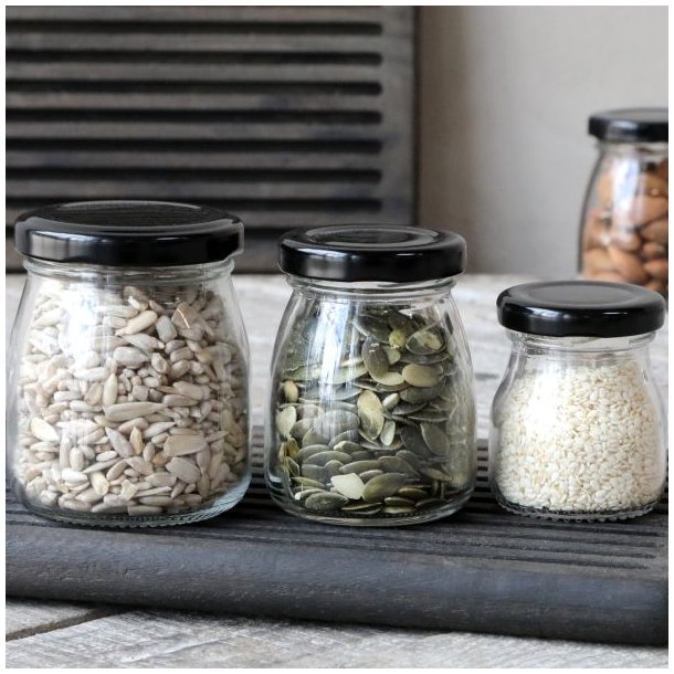 Opbevaringsglas med sorte l�g fyldt med solsikkefr�, gr�skarkerner og sesamfr� p� en m�rk tr�bakke.