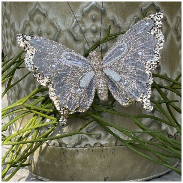 Champagnefarvet organza sommerfugl med glimmer og clips fastgjort p� gr�nne grene foran en m�nstret urtepotte.