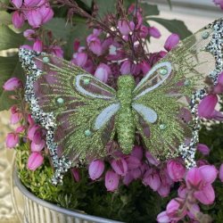 Gr�n organza sommerfugl med clips, hvide aftegninger og similisten sat p� en potteplante med sm� lyser�de blomster.