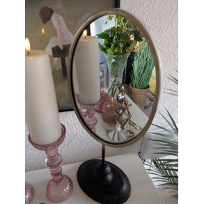 Et elegant ovalt bordspejl med en spinkel guld- eller messingfarvet ramme st�r p� en sort fod. I spejling ses en stemningsfuld styling med to h�je hvide bloklys i lyser�de glasholdere med struktur, dekorative balletdanser-figurer i messingfinish og en keramikvase med friske gr�nne grene og blade. I baggrunden til h�jre anes en potteplante i en kurv og til venstre et indrammet kunstv�rk.