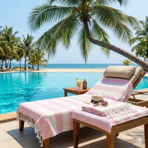 Et kvadratisk milj�billede af et dr�mmende ferieparadis med en luksuri�s solvogn ved en infinity-pool og en sandstrand. Et pink- og hvidstribet hamam-badeh�ndkl�de fra La Vida er lagt flot p� solvognen. En hat og en forfriskende drink st�r ved siden af. I baggrunden ses palmetr�er og det turkisbl� hav under en skyfri himmel.