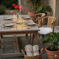 plaider-bomuld-fletkurv-terrasse-miljoe Et stemningsfuldt udend�rs milj�billede, hvor to m�nstrede bomuldsplaider i d�mpede beige og gr� nuancer ligger sirligt rullet sammen med et brunt b�nd og en tr�knap nede i en rund fletkurv med hank. Kurven st�r p� fliserne ved siden af et rustikt tr�bord og en krukke med en gr�n Monstera-plante.