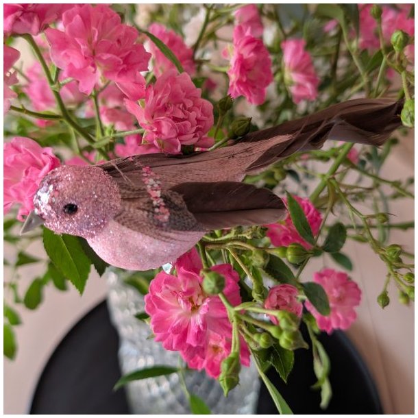 Sidel�ns n�rbillede af en 15 cm lang rosabrun dekorationsfugl med m�rke naturfjer og glitrende detaljer i en rosenbuket.