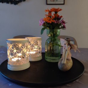 To r�hvide metalfyrfadsstager med udsk�ret m�nster og patina placeret p� et sort fad ved siden af en gr�n glasvase og en lille fe-figur.