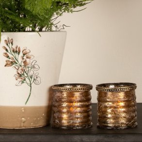 Kvadratisk milj�billede af to t�ndte, ravfarvede glasfyrfadsstager med rillem�nster og kant med perledetaljer, st�ende ved siden af en hvid urtepotteskjuler med blomstermotiv p� en m�rk hylde.