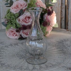 En elegant, rillet glasvase p� fod med en timeglasformet krop og en b�lget kant foroven. Vasen st�r tom p� et dekorativt, udsk�ret bord med en m�nstret overflade, og i baggrunden ses en buket af lyser�de roser og eucalyptus, mod en rustik tr�v�g.