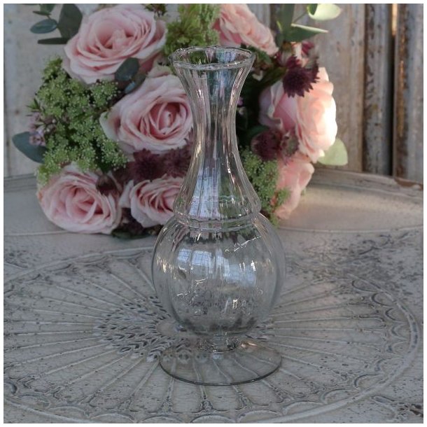 En elegant, rillet glasvase p� fod med en timeglasformet krop og en b�lget kant foroven. Vasen st�r tom p� et dekorativt, udsk�ret bord med en m�nstret overflade, og i baggrunden ses en buket af lyser�de roser og eucalyptus, mod en rustik tr�v�g.