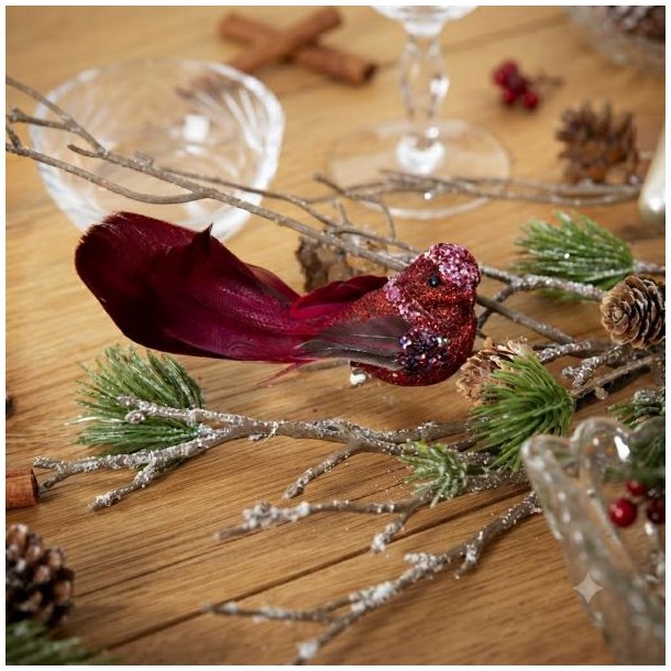 R�d dekorationsfugl med glimmer placeret p� en gren i en hyggelig juledekoration med grankogler og kanelst�nger p� et tr�bord.