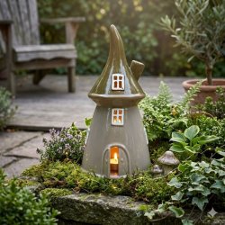 Eventyrligt keramikhus med gr�nt tag placeret i haven med t�ndt lys. Skovhuset er omgivet af planter og mos p� en terrasse, hvilket skaber en hyggelig stemning som fyrfadsstage.