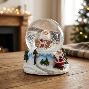 Stemningsfuldt milj�billede af en dekorativ snekugle med julemand og snefnug placeret p� en tr�bordplade med julelys i baggrunden.