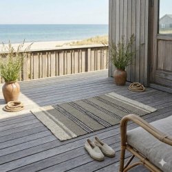 Rustikt s�gr�st�ppe med striber i m�lene 120x180 cm placeret p� en tr�terrasse. I baggrunden ses en smuk havudsigt med strand og klitter. T�ppet er stylet med et par hvide espadriller, rustikke krukker med marehalm, reb og en kurvestol, hvilket skaber en hyggelig og nordisk sommerhusstemning.