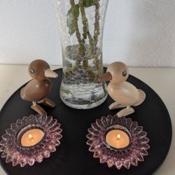 sort-traefad-la-vida-miljoe-traefugle-solsikkestager Det runde sorte tr�fad fra La Vida er pyntet med to sm� dekorative tr�fugle i m�rkt og lyst tr�, der st�r p� hver sin side af en klar glasvase med blomster, foran to t�ndte lysestager i lyser�dt glas formet som solsikker.