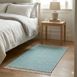 Et rektangul�rt, v�vet bomuldst�ppe i en flot turkisbl� farve med fine striber og lyse frynser i enderne. T�ppet m�ler 150x70 cm og er placeret p� et lyst tr�gulv ved siden af en redt seng med lyst senget�j. Ved siden af sengen st�r et enkelt sidebord i lyst tr� med en lampe og et glas vand, hvilket skaber en rolig og moderne skandinavisk atmosf�re i sovev�relset.