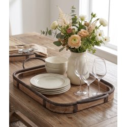 Firkantet milj�billede af den store rektangul�re tr�bakke med m�rk jernkant og h�ndtag, placeret p� et spisebord af tr�. Bakken er d�kket op med en stabel keramiktallerkener, sk�le, fine vinglas og en hvid vase med friske blomster.