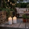 Et stemningsfuldt milj�billede, der viser to t�ndte udend�rs LED bloklys p� en vejrbit tr�terrasse i skumringen, med en stenmur og en hyggelig loungeplads i baggrunden.