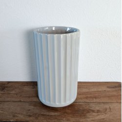 Den h�je lysegr� vase st�r placeret p� et rustikt tr�bord i en hyggelig indend�rs opstilling. Vasens rillede overflade fanger lyset smukt.