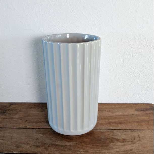 Den h�je lysegr� vase st�r placeret p� et rustikt tr�bord i en hyggelig indend�rs opstilling. Vasens rillede overflade fanger lyset smukt.