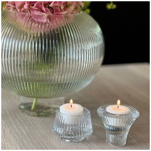 To runde og kegleformede vendbare fyrfadsstager i klart krystalglas opstillet med t�ndte fyrfadslys ved en blomstervase.