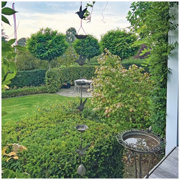 Et h�jt vindspil i antik brunt jern fotograferet i fuld figur i en gr�n have, hvor det h�nger dekorativt mellem buske og tr�er.