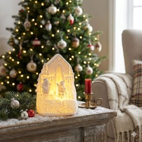 Det m�nstrede hvide ceramic-vinterhus med t�ndte LED-lys (fra produktbilledet) st�r som centerpiece i et julemilj� p� en sne-d�kket kaminhylde med granris og en brass-lysestage. I baggrunden ses et juletr� med lys.