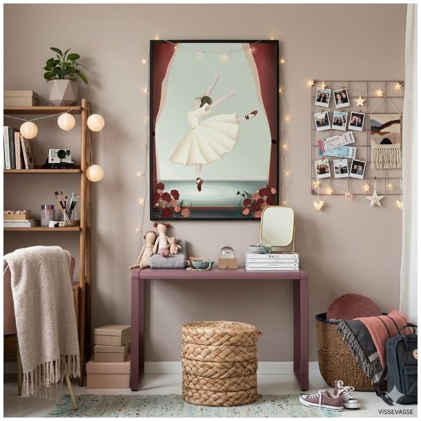 Et hyggeligt 1:1 milj�billede fra et teenagev�relse, hvor en indrammet Vissevasse plakat med en dansende ballerina i en lys kjole h�nger p� en st�vet rosa v�g over et rosa sminkebord. Plakaten er dekoreret med en fin lysk�de langs rammen. P� bordet findes sm� stofdukker, kosmetik og et spejl, og p� gulvet foran st�r en flettet skammel samt en kurv med t�pper. Til h�jre p� v�ggen h�nger et opslagsv�rk i metal med personlige billeder, hvilket skaber en personlig og varm atmosf�re under et bl�dt dagslys.