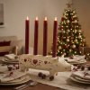 Stemningsfuldt milj�billede af Wilma Gris adventsstage med r�de lys placeret p� et d�kket spisebord med et pyntet juletr� i baggrunden.