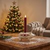 Stemningsfuldt milj�billede af den lille Wilma Gris lysestage i hvid polyresin med r�dt lys, st�ende p� et tr�bord i stuen med et pyntet juletr� i baggrunden.