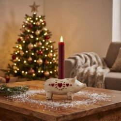 Stemningsfuldt milj�billede af den lille Wilma Gris lysestage i hvid polyresin med r�dt lys, st�ende p� et tr�bord i stuen med et pyntet juletr� i baggrunden.