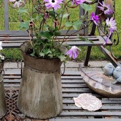 Et n�rbillede af den store (H20,5 cm) meleret zinkurtepotte p� haveb�nken. Billedet fokuserer p� potten med den lilla blomst, metalhanken og de detaljerede m�nstre p� metalb�nken. Baggrunden er sl�ret, men viser en lille fugleskulptur.