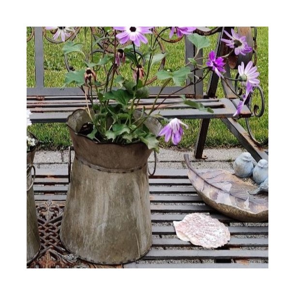 Et n�rbillede af den store (H20,5 cm) meleret zinkurtepotte p� haveb�nken. Billedet fokuserer p� potten med den lilla blomst, metalhanken og de detaljerede m�nstre p� metalb�nken. Baggrunden er sl�ret, men viser en lille fugleskulptur.