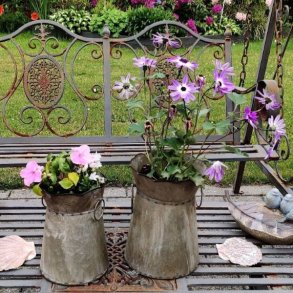 Et havemilj�billede af de to meleret zinkurtepotter, placeret sammen p� en rustik metalhaveb�nk. Den store potte (H20,5 cm) rummer en plante med lilla blomster, og den mellemste potte (H19 cm) indeholder lyser�de flittiglise. Baggrunden viser en frodig have med rhododendron i blomst.