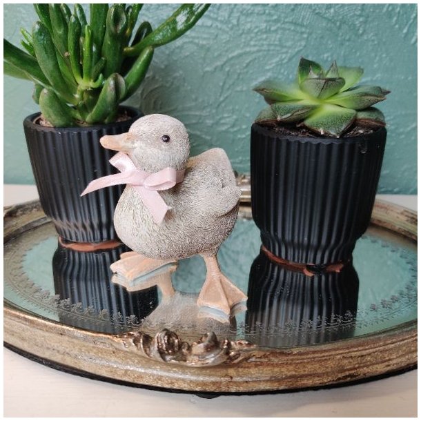 �llingefigur med et lyser�dt b�nd om halsen, st�ende p� en dekorativ spejlplade med to sm� sukkulenter i sorte krukker.