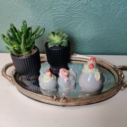 Tre gr� p�skefigurer af h�ns med t�rkl�der i pastelfarver, placeret p� en oval spejlbakke med guldkant ved siden af to sm� gr�nne planter i sorte krukker.  De sm� figurer m�ler ca. 4 cm i h�jden, mens den st�rste h�ne m�ler ca. 7 cm.