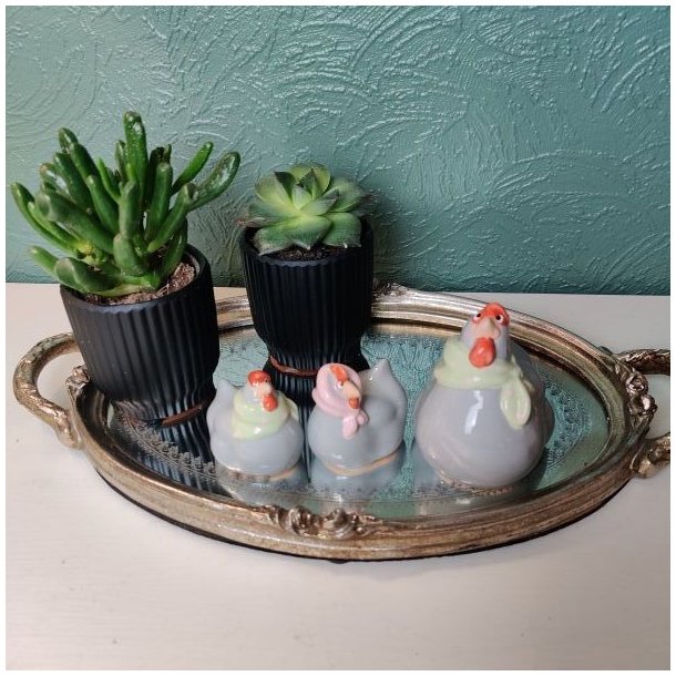 Tre gr� p�skefigurer af h�ns med t�rkl�der i pastelfarver, placeret p� en oval spejlbakke med guldkant ved siden af to sm� gr�nne planter i sorte krukker.  De sm� figurer m�ler ca. 4 cm i h�jden, mens den st�rste h�ne m�ler ca. 7 cm.